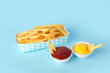 Paper box with tasty french fries and bowls of sauces on blue background