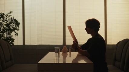 Silhouette of adult male sitting at the bistro bar, man having lunch alone, holding and reading...