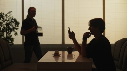 Silhouette of man sitting at the fast food bar holding reading social media on smartphone, waitress coming at the back.
