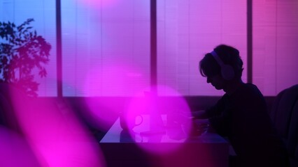 Silhouette of male sitting at the food bar at evening in neon light, wearing headphones, working on...