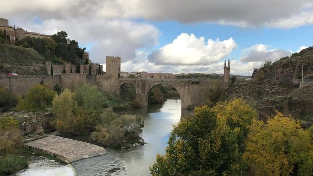 Panorama shot of Alcantara arch bridge over Tagus river, reflecting cotton clouds and blue sky. Reveals Alcantra royal palace on hill. Toledo, Spain.
