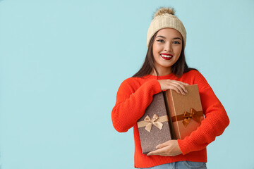 Beautiful young Asian woman in warm hat with gift boxes on blue background