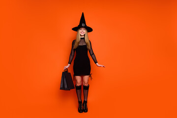 Full size photo of pretty blonde teenager girl stand hold shopping bags wear black halloween outfit isolated on orange color background
