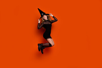 Fototapeta na wymiar Full size photo of pretty blonde teenager girl jumping cheerful have fun dressed black halloween outfit isolated on orange color background