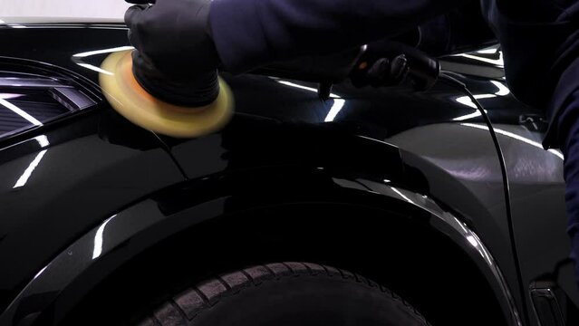 Close-up of a car mechanic polishing a black car with wax and a polishing machine in a modern car dealership. 