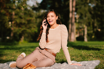 Young woman is enjoying a picnic in the park and talking on a smartphone