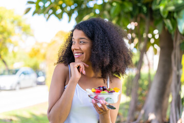 Young African American woman holding a bowl of fruit at outdoors thinking an idea and looking side