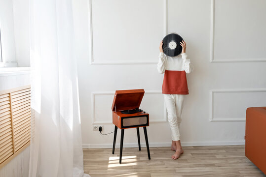 Retro vinyl record music modern trend. Young tennage boy with vinyl record in hands, hiding face behind record, standing near wooden brown turntable. White wall background, home interior, lifestyle