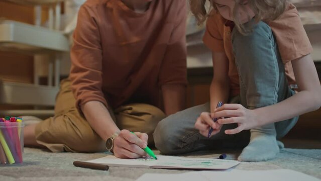 Medium shot of caucasian woman sitting on floor with her son and helping him with drawing picture using marker pens