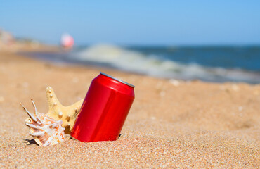 Red aluminum can with a drink and shells on the beach near the sea.
