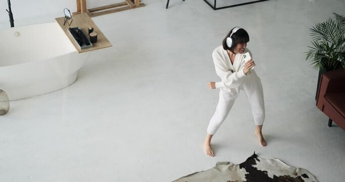 Carefree Caucasian woman, donning headphones and holding phone, dances and sings with abandon. Her joyful movements and melodious voice create a scene of unbridled happiness.