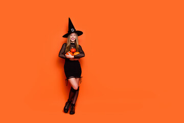 Full body photo of attractive blonde teen woman hold pumpkins dressed black halloween clothes isolated on orange color background