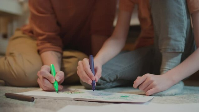 Selective focus medium close-up shot of unrecognizable mother and son sitting on floor in bedroom drawing colorful pictures with marker pens