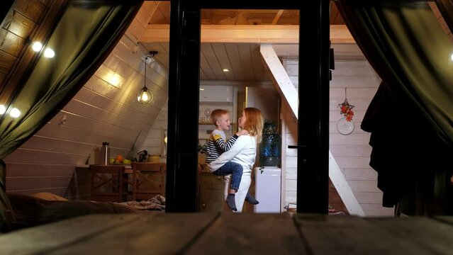 Mother and son are having fun and dancing in a one-story wooden bungalow house with lanterns in the evening and a triangular roof descending to the ground. Winter vacation in the forest.