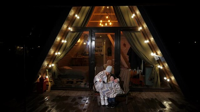 Mother and son covered themselves with a blanket and sit in an armchair near a small triangular wooden house in the evening. The house is a bungalow with lanterns and a roof descending to the ground.
