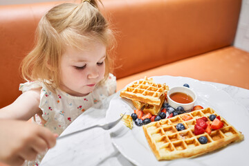 Happy 3 year old girl eat. Breakfast in cafe. Classic Viennese waffles with ice cream, berries and Maple syrup. Table in the restaurant.