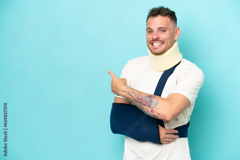 Wall mural young caucasian man wearing a sling and neck lace isolated on blue background pointing back