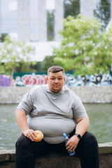 Naklejka na ściany i meble fat man eating a burger and drinking water after exercising in the park