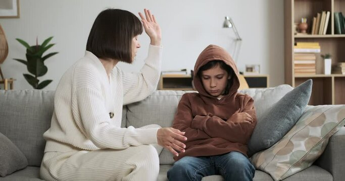 Furious and upset Caucasian mother raises voice at misbehaving son at sofa in the living room. Tensions run high as they navigate a moment of conflict.