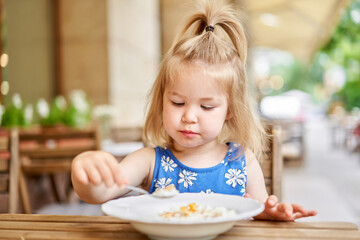 Little kid having breakfast at cafe. Adorable girl drinking still water, eating rice porridge with...