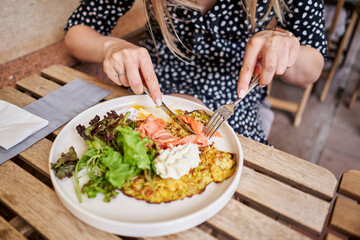 Woman cuts Zucchini pancakes, with avocado, cheese cream, salmon and egg . Healthy breakfast, protein. Restaurant dish. Breakfast in cafe on a sunny morning, on the summer veranda