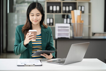 Asian businesswoman sits in a office working on laptop and enjoys a coffee.