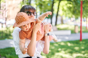 Portraits young mother and her daughter of acute 10 months old baby girl. Walking on the playground, on a sunny summer day.