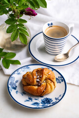 Homemade traditional Swedish cinnamon bun (swirl) on white and blue porcelain plate and cup of espresso coffee on white background.