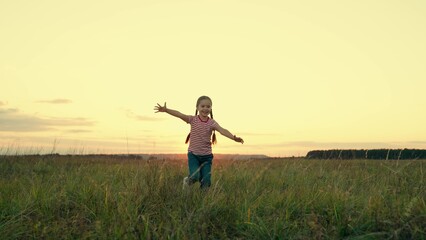 Fototapeta na wymiar Positive little girl runs spreading arms for embrace in country field at sunset