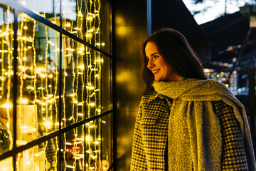 Cheerful woman looking at a shop window with Christmas lights from the street at night