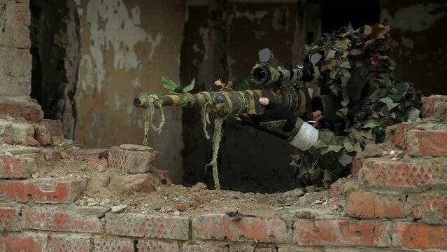 Portrait of a military sniper in camouflage in a destroyed brick building, he takes aim standing near the window. Military operation. The sniper shoots at the enemy.