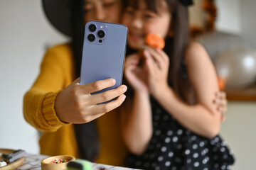 Smiling asian mother taking a selfie on smart phone and having fun with her daughter in Halloween decorated kitchen