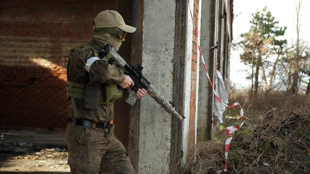 A man in a military camouflage uniform with an automatic rifle with an optical sight takes aim while standing in a destroyed brick building. Attack on the battlefield. Airsoft.