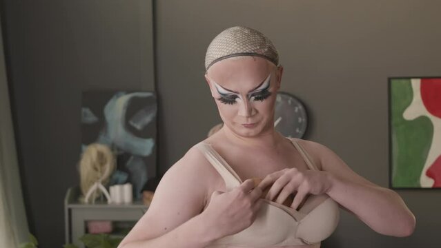 Waist up of Caucasian queer man with impressive drag makeup using pushup bra pads to make fake breasts while getting dressed for drag show at home