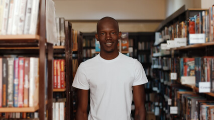 Fototapeta na wymiar Black Man In Library Turns And Looks At Camera Smiles, Standing Between The