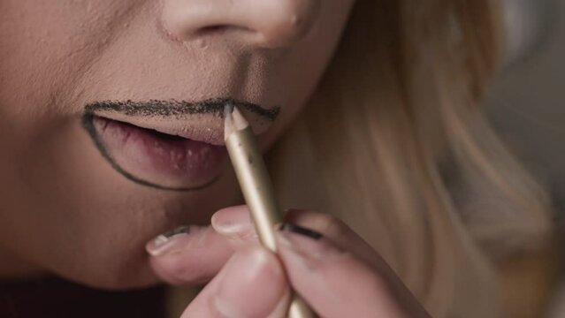 Closeup of unrecognizable queer man applying brown lip liner outside of natural lip contour while doing drag makeup in backstage room