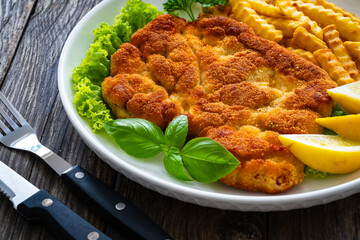 Crispy breaded seared chicken cutlet with French fries,   lemon and fresh vegetables on wooden table
