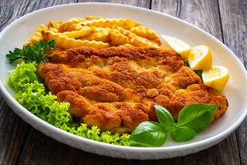 Crispy breaded seared chicken cutlet with French fries,   lemon and fresh vegetables on wooden table
