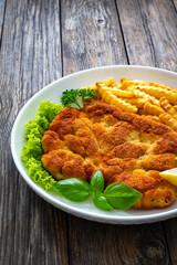 Crispy breaded seared chicken cutlet with French fries,   lemon and fresh vegetables on wooden table
