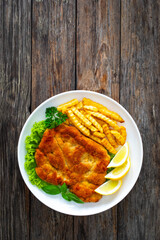 Crispy breaded seared chicken cutlet with French fries,   lemon and fresh vegetables on wooden table
