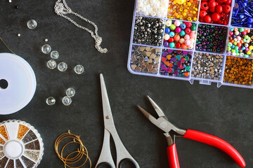 Box with colorful beads, string, wire, chain, scissors, pliers and hammer on dark background. Various jewelry making supplies. Top view.