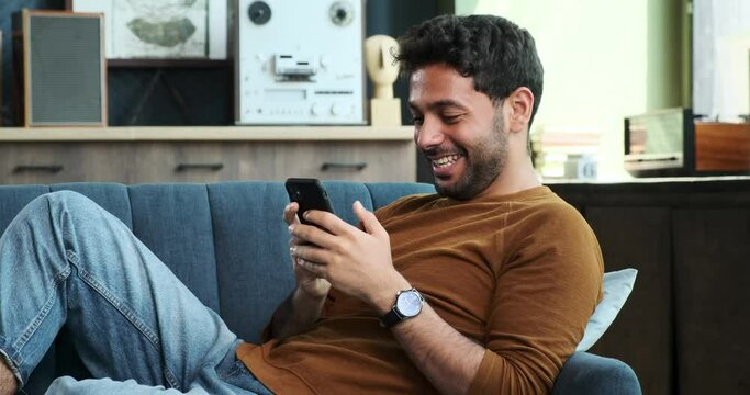 Middle Eastern man comfortably sits on a sofa in the living room, using phone. His relaxed demeanor showcases the convenience of staying connected and productive while in a home setting.