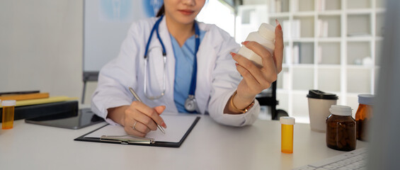 Female doctor writing in a notebook and holding a medicine bottle works at a computer while there were many medicine bottles on the table while sitting at a work desk