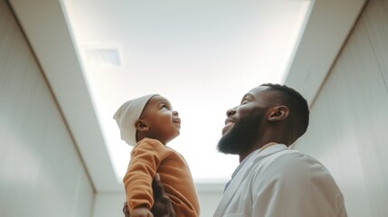 Bottom view of an African American male doctor wearing medical clothes talks to a child in a hospital corridor. - obrazy, fototapety, plakaty