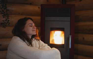 A young woman wraps herself in a warm blanket while sitting by a pellet stove in a wooden log...