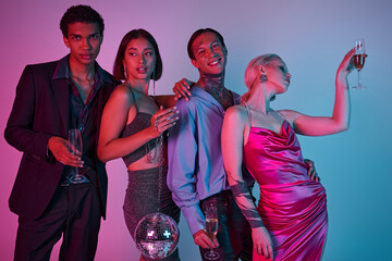 happy interracial friends holding champagne glasses and disco ball on purple pink, Christmas party