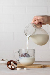 A hand pours milk from a jug into a glass cup with cocoa, chocolate bomb. Making hot chocolate. Vertical frame, light background.