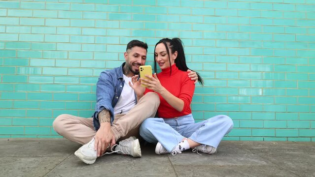 young couple sitting on the floor while visiting the city of london, tourists taking selfies with smart phone, on turquoise wall.