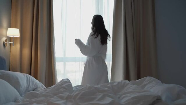Happy active woman experiencing euphoria of pleasure, carefree dancing in the morning in front of a full-length panoramic window of a bedroom in a hotel apartment