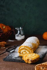 Dessert, a whole pumpkin sponge roll with cream cheese cream, sprinkled with powdered sugar, on a black slate board on a brown concrete background. Pumpkin recipes.